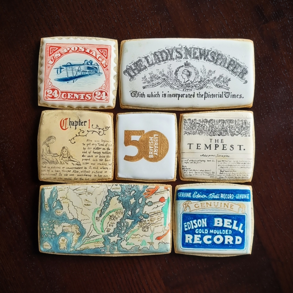 A set of 7 biscuits arranged in a square. In the top left is a biscuit version of the 1918 ‘inverted Jenny’ stamp. The royal icing has a scalloped edge, and the words ‘U.S. POSTAGE … 24 CENTS’ frame a navy-blue, upside-down image of a biplane. The top-right biscuit reads ‘The Lady’s Newspaper’ in ornate black lettering above a cameo of Queen Victoria. Below that is a biscuit depicting the title page of ‘The Tempest’ from Shakespeare’s First Folio. The bottom-right biscuit is inspired by a wax cylinder label: it reads ‘Genuine Edison Bell Gold Moulded Record’ in a vintage typeface. To the left is a large biscuit painted to look like a section of the Cotton Mappa Mundi, with patchy blue seas, red rivers, and turquoise mountain ranges. In the centre-left is a biscuit based on Lewis Carroll’s ‘Alice’s Adventures Under Ground’. ‘Chapter 1’ is written in a Gothic font above a hand-drawn illustration of Alice and her sister. The central biscuit features the British Library at 50 logo in gold.