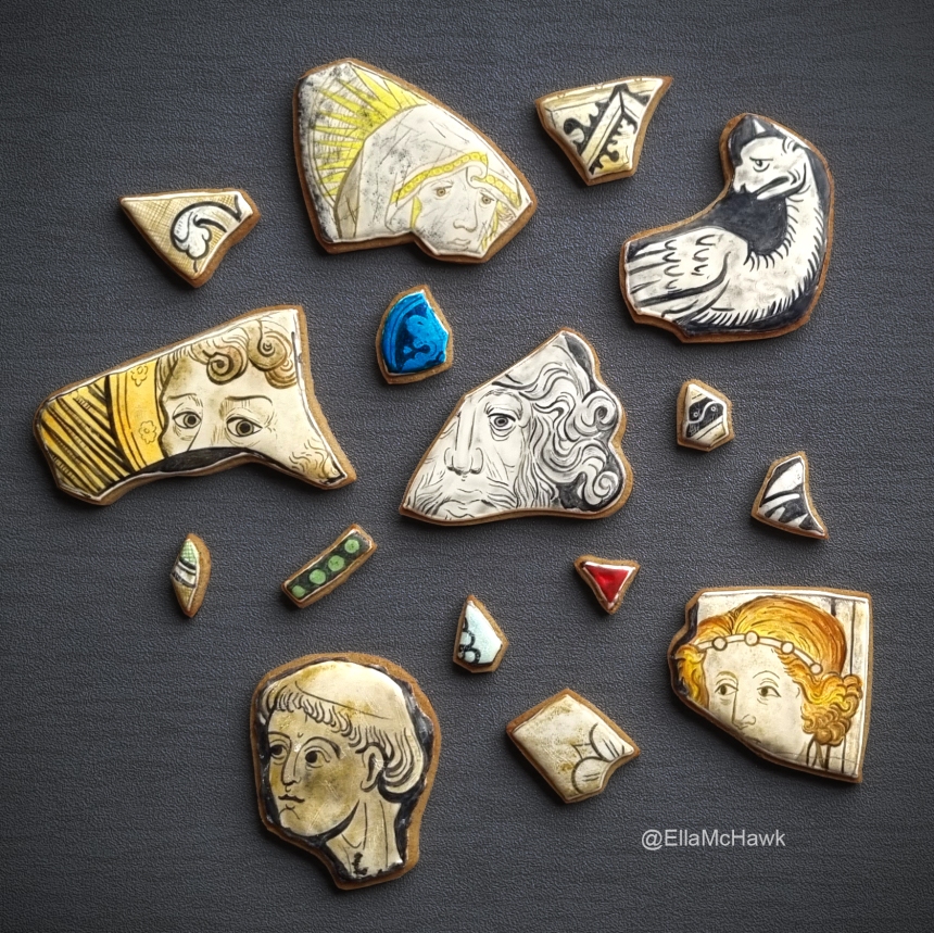 A collection of biscuits made to look like stained glass window fragments. There are six larger biscuits featuring different faces: a woman looking downwards with sad eyes; a griffin-like creature with wings and a long neck; an old man with long facial hair; a woman with golden hair and a simple tiara; a bald man painted in monochrome; a man with mournful eyes, curled hair, and a vivid yellow background. The larger biscuits are surrounded by a scattering of smaller fragments. There is a bright blue semi-circular shard painted with a leaf pattern, and a jewel-red fragment in a triangular shape. Other small biscuits have bold black-and-white patterns made up lines, circles, and floral shapes. Many of the biscuits are made to look dirty and scratched. 