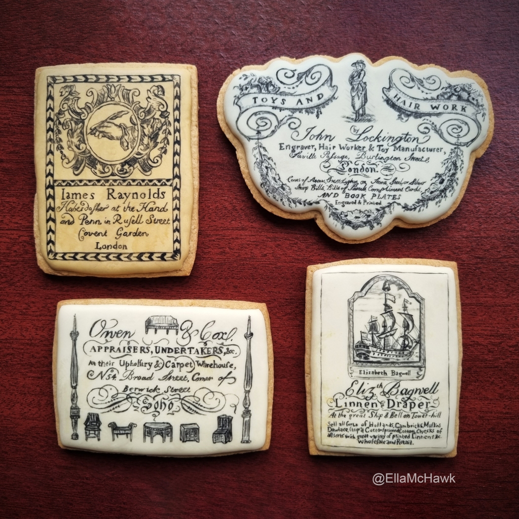 A set of four biscuits against a maroon wooden background. The biscuits are iced in different shades of cream. Three are rectangular, and one has a cloud-like shape. Each biscuit is painted to look like an eighteenth-century printed trade card. One has ‘James Reynolds’ written in large type, spelt with an I instead of a J. Beneath, in a curly calligraphic font, the biscuit reads: ‘Haberdasher at the Hand and Pen in Russell Street, Covent Garden, London’. The design is finished with an extravagantly-framed hand holding a quill. The other biscuits read: ‘John Lockington: Engraver, Hair Worker & Toy Manufacturer’; ‘Elizabeth Bagwell, Linen Draper at the great Ship & Bell on Tower Hill’; and ‘Owen & Cox, Appraisers, undertakers, etc.’ Elizabeth Bagwell’s card features an image of an eighteenth-century ship. Owen & Cox’s design is framed with tiny line drawings of furniture pieces. 