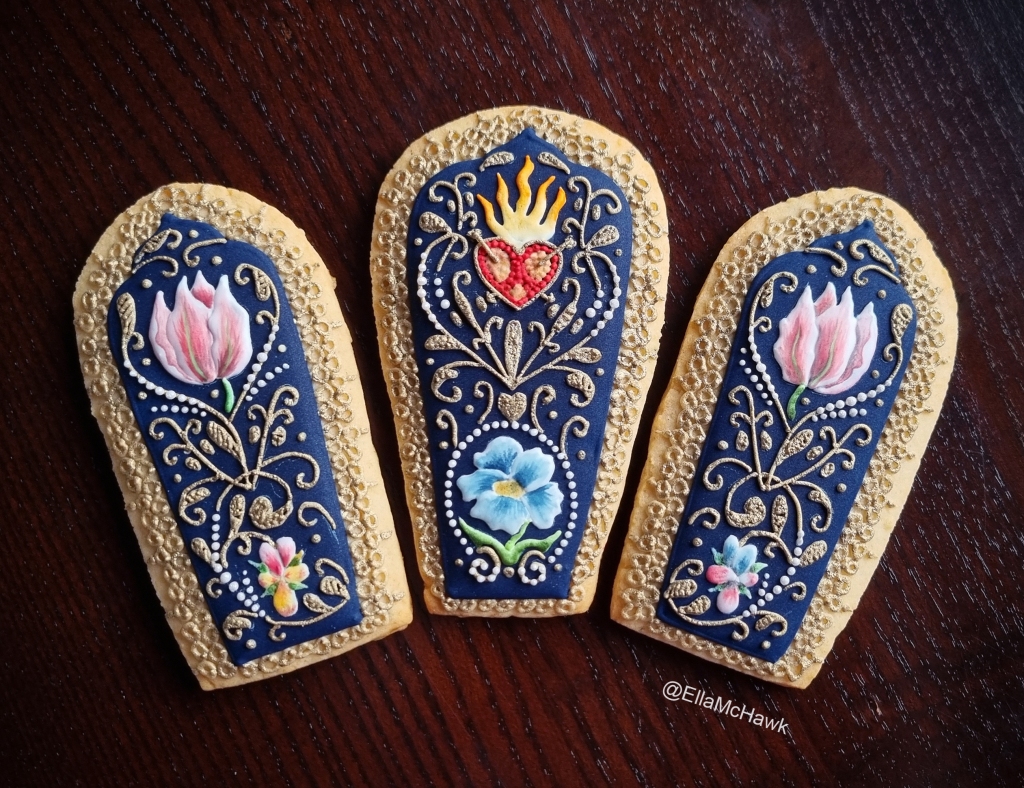 A set of three petal-shaped biscuits arranged into a fan against a dark wooden background. The biscuits are covered with a deep, rich blue icing and embellished with intricate details. At the centre of the arrangement is a textured red heart with bright yellow flames, pierced with two tiny gold arrows. The heart is surrounded by curling gold vines, delicate pearl details, and hand-painted flowers. There is a large blue pansy, two pink tulip-like flowers, and two smaller pansies in shades of pink, yellow, and blue. Each biscuit is edged with gold lace made up of small icing circles. 