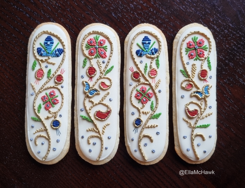 Four narrow biscuits with rounded ends laid on a dark wooden background. A golden vine curves up the centre of each ivory biscuit, sprouting colourful flowers and leaves. The flowers are piped in royal icing to look like the texture of embroidery. The ivory background of each background is spotted with small silver details that resemble metal sequins. 